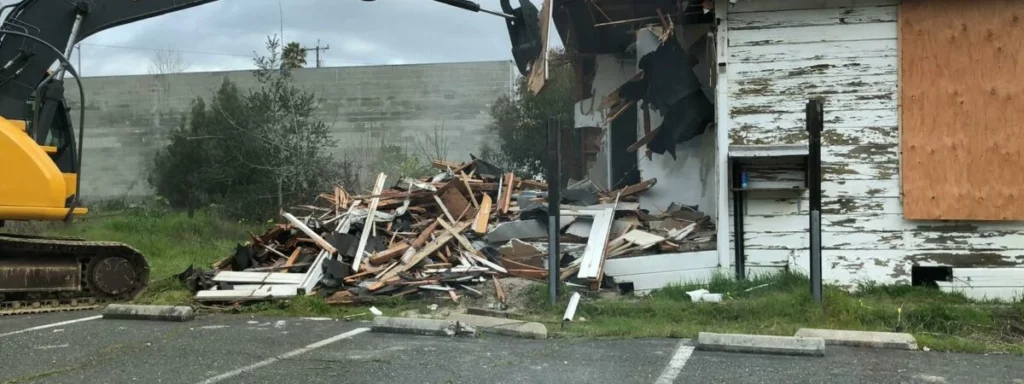 A yellow excavator demolishes an old, weathered building in Marin County. Debris and wooden planks are scattered on the ground next to the partially collapsed structure, set against the picturesque backdrop of the Bay Area near San Francisco.