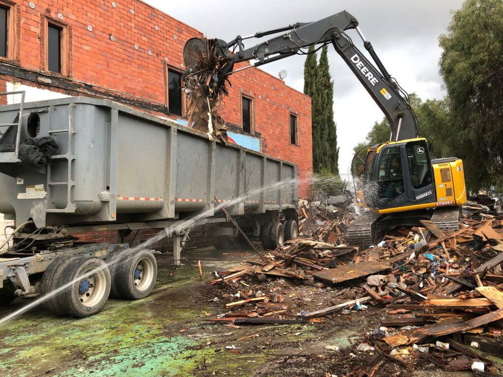 A construction site in Oakland shows a backhoe loader dumping debris into a large dumpster truck. Wood and other debris are scattered around, and a hose sprays water on the scene.