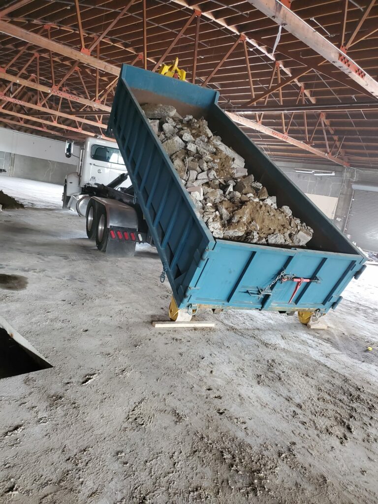 A blue dump truck is unloading rubble in a large, unfinished building with exposed metal beams and a concrete floor in Oakland.