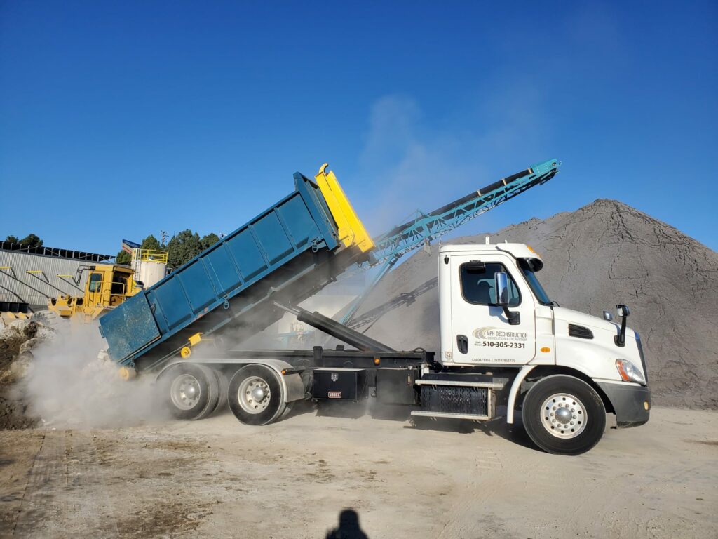 A dump truck unloading a load of grey gravel at a construction site in Palo Alto creates dust clouds under the bright blue sky.