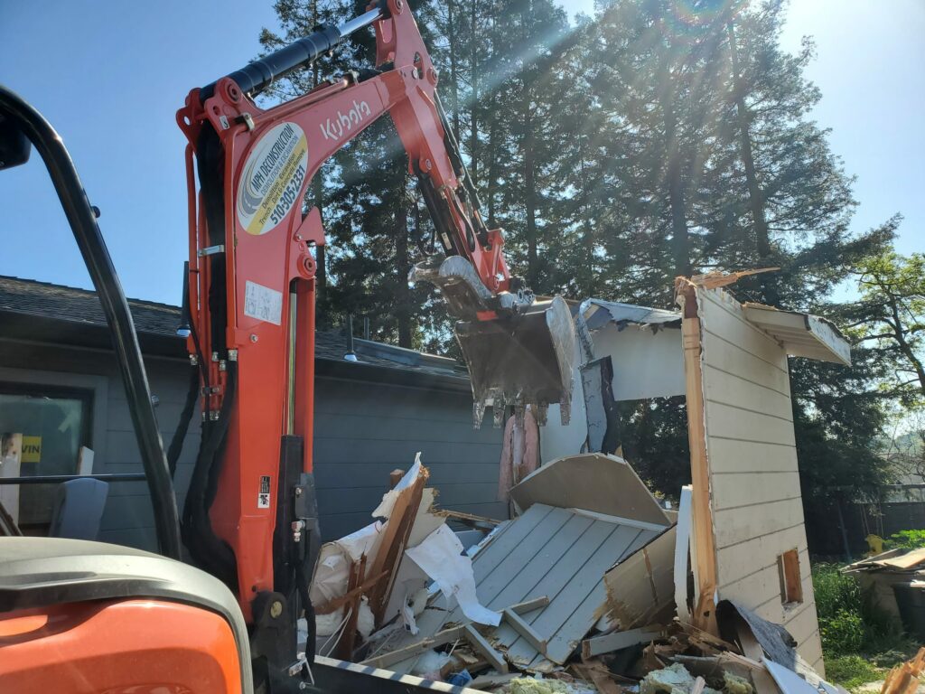 An orange excavator demolishes a small wooden shed, with its arm tearing through the walls in a quiet corner of Marin County. Sunlight filters through the trees in the background, casting dappled shadows on the scene. Residential Demolition