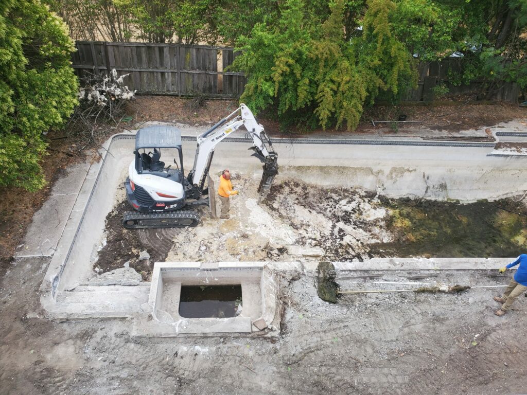 A construction worker in an orange shirt operates a small excavator to dig and remove debris from an empty swimming pool surrounded by greenery in the Bay Area.