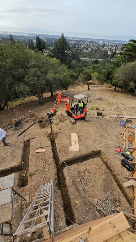 A small excavator performs excavation work on a hillside construction site in the Bay Area, with workers, building materials, and a city skyline visible in the background.