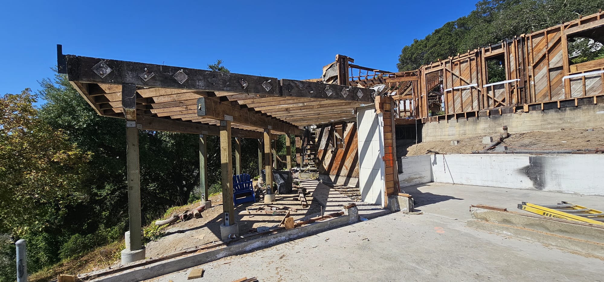 A construction site featuring a partially built structure with exposed wooden beams and supports. Trees and clear blue sky are visible in the background.