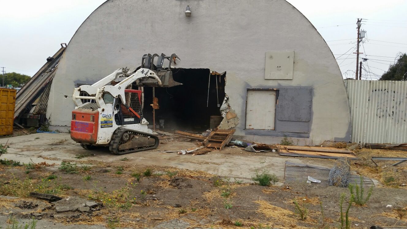 A small bulldozer from a professional demolition company efficiently dismantles a large, arched warehouse. Debris and dirt envelop the area.