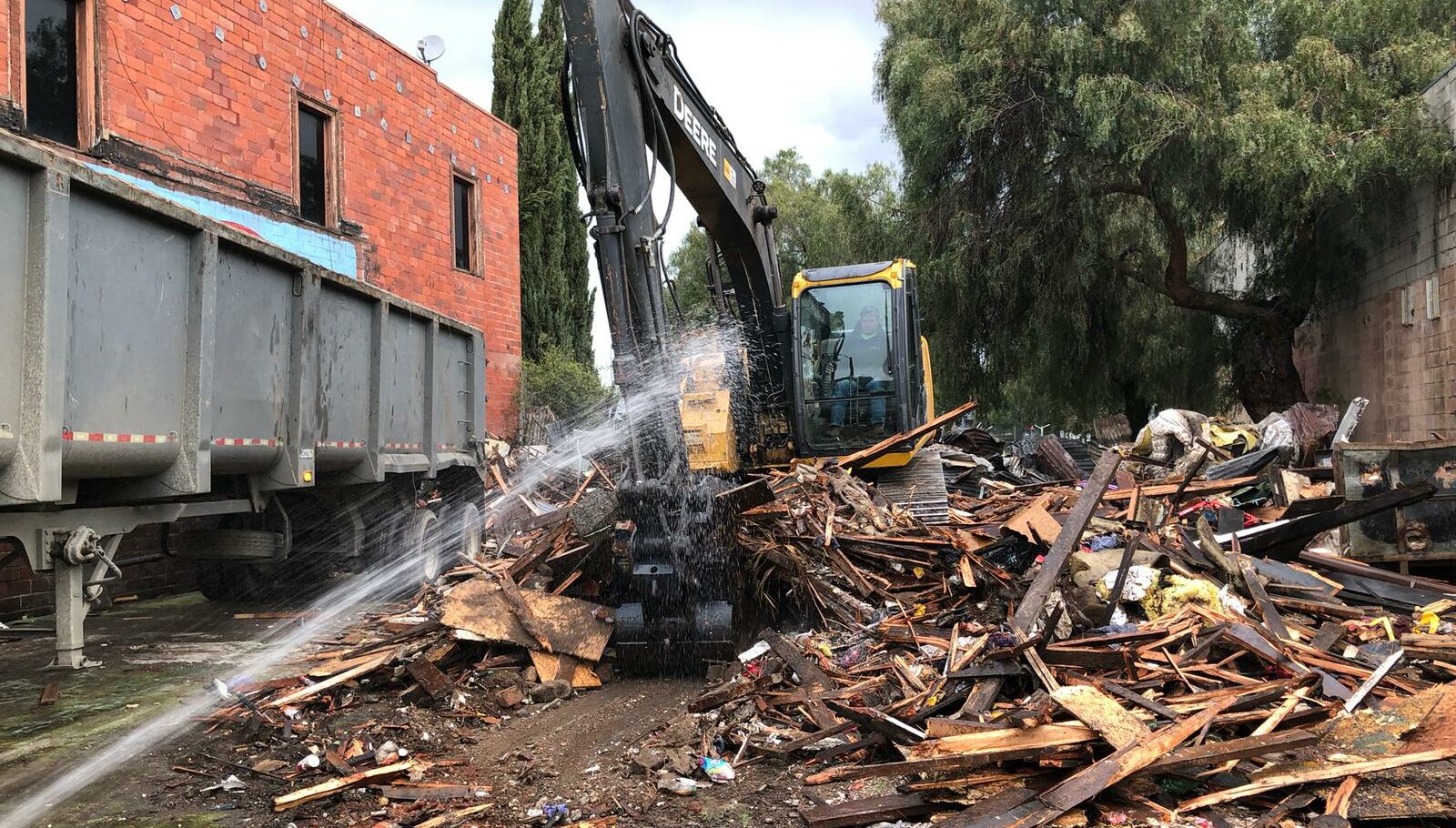 An excavator clears debris while water is sprayed over a demolition site next to a brick building.