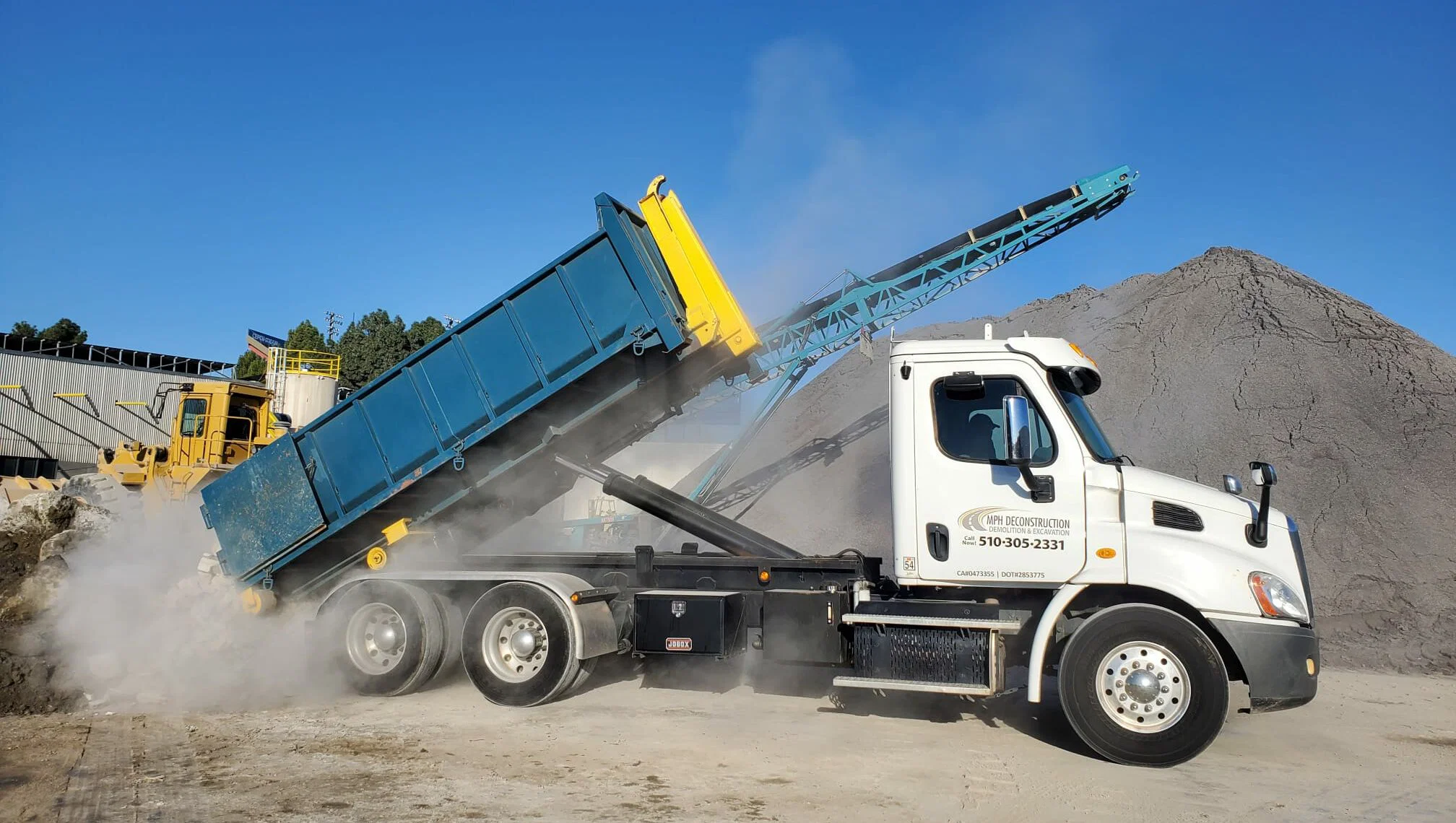 A dump truck unloading gravel at a construction site, with dust rising around the truck bed.