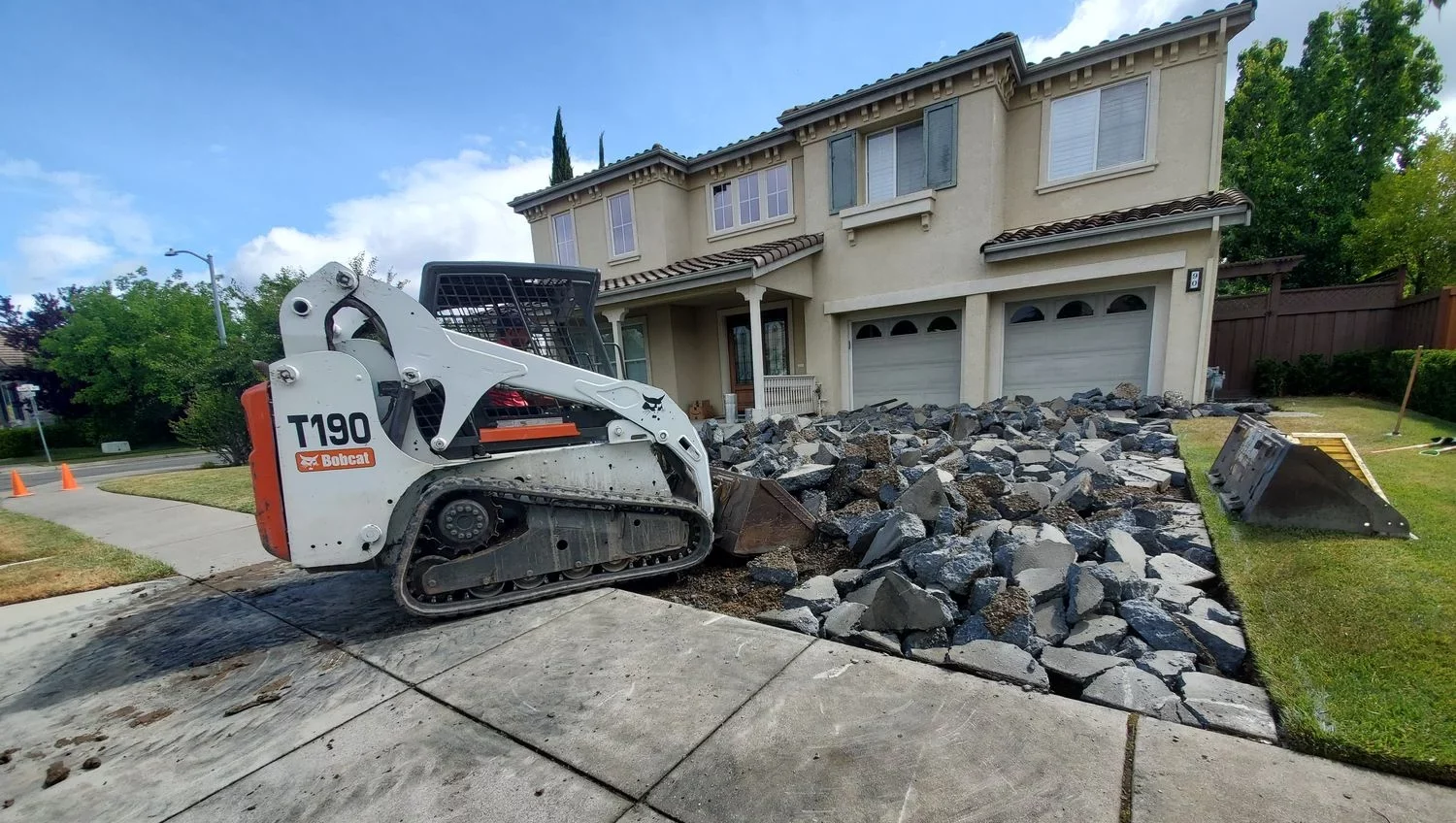 A skid steer loader breaks up a concrete driveway in front of a two-story suburban house.