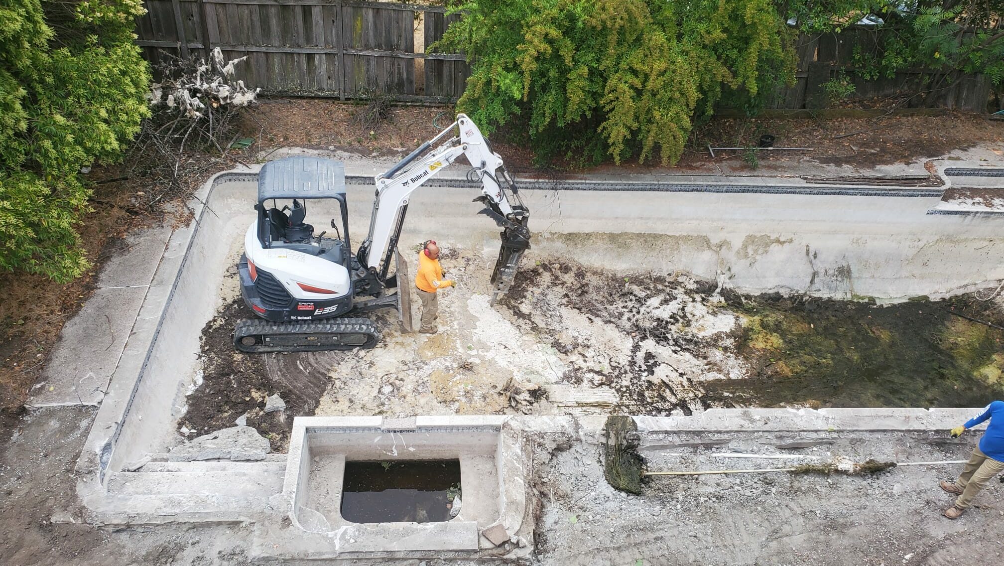 A worker uses a small excavator to break up concrete in an empty, abandoned swimming pool.