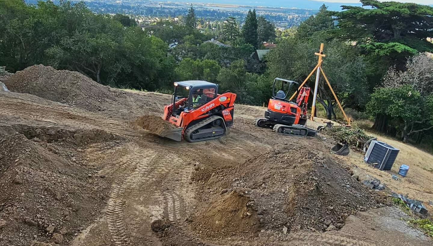 Two orange excavators moving dirt on a construction site with trees and a cityscape in the background.