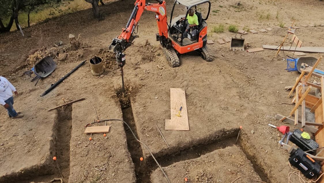 A small excavator digs foundation trenches at a construction site on a hill overlooking a city.